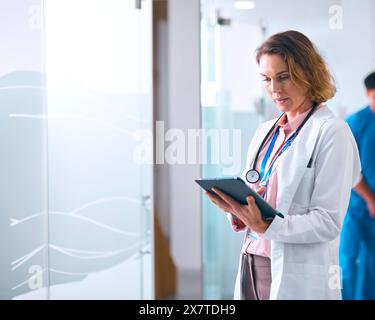 Médecin féminin en manteau blanc avec tablette numérique à l'hôpital avec des collègues en arrière-plan Banque D'Images