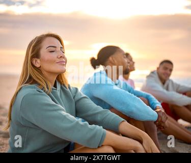 Femme avec les yeux fermés se relaxant avec des amis en vacances assis sur la plage regardant le lever du soleil ensemble Banque D'Images