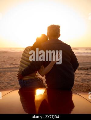 Vue arrière du couple senior retraité assis sur le capot de la voiture de sport classique à la plage regardant le lever du soleil Banque D'Images