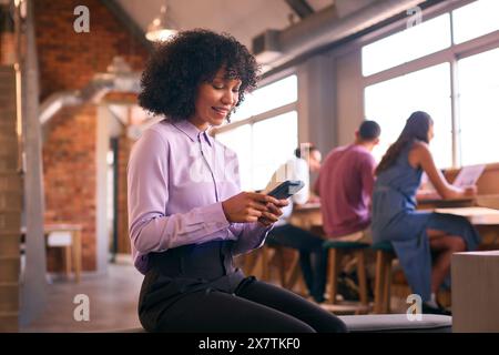 Femme d'affaires souriante avec téléphone portable travaillant dans le bureau Open Plan avec des collègues en arrière-plan Banque D'Images