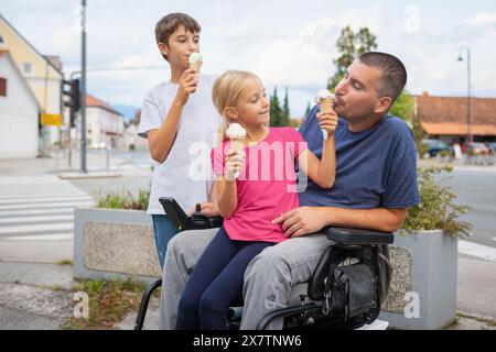 Père handicapé en fauteuil roulant s'amusant avec ses enfants mangeant de la crème glacée. Concepts de famille et de déficience physique. Banque D'Images
