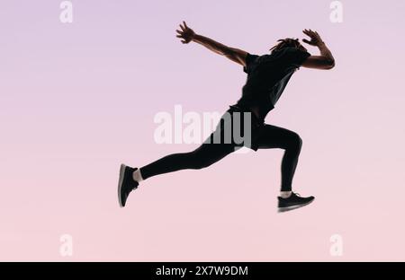 Fit Man avec dreadlocks saute en plein air, mettant en valeur son dévouement pour le fitness. Sur fond rose, il pratique la course à pied et les sprints en sportswear Banque D'Images