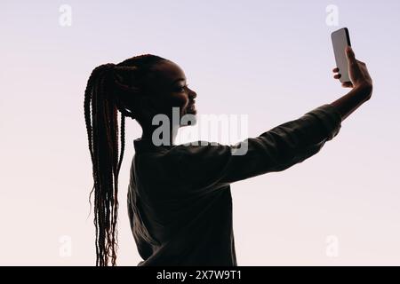 Une femme souriante d'une vingtaine d'années se tient dans un studio pour un autoportrait. Elle tient un téléphone portable et prend un selfie, soulignant sa confiance en soi et sa vie Banque D'Images