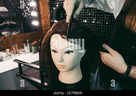 Coiffeur pratique de peignage sur mannequin. Homme jeune coiffeur diligemment avec enthousiasme avec la longue barbe et les cheveux dans le gilet, coiffure de peignage pratiquant sur mannequin dans le salon de beauté .concept pratique compétences école de formation coiffeurs Banque D'Images