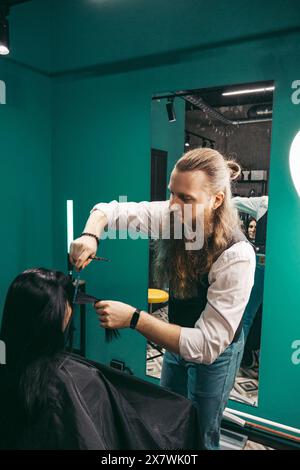 Coiffeur fait une coiffure pour femme. Homme coiffeur avec une longue barbe dans un gilet, fait habilement un coiffure avec un peigne et un ciseau de coupe de cheveux à une femme avec de longs cheveux noirs dans la boutique d'un coiffeur .concept de compétences approche client. prise de vue moyenne Banque D'Images