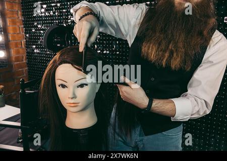 Coiffeur pratique de peignage sur mannequin. Homme jeune coiffeur diligemment avec enthousiasme avec la longue barbe et les cheveux dans le gilet, coiffure de peignage pratiquant sur mannequin dans le salon de beauté .concept pratique compétences école de formation coiffeurs Banque D'Images