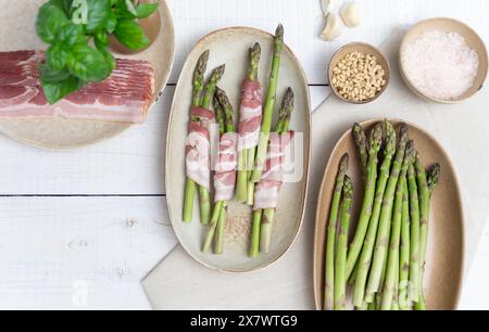 Asperges vertes croquantes montrées sur des assiettes beiges, prêtes pour le four. Encadré par des assiettes avec du bacon et d'autres ingrédients comme l'ail et les pignons de pin Banque D'Images