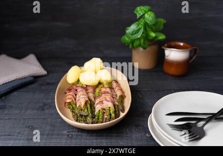 Asperges vertes cuites cuites enveloppées dans du bacon et servies avec des pommes de terre bouillies sur de belles assiettes beiges sur fond sombre Banque D'Images