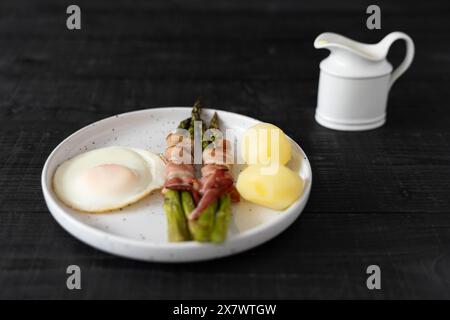 Asperges vertes cuites au four enveloppées dans du bacon et servies avec des pommes de terre bouillies et des œufs, sur de belles assiettes blanches sur une surface en bois noir Banque D'Images