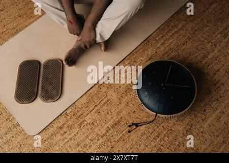 Vue de dessus photo recadrée d'un jeune homme africain méconnaissable en transe méditant dans une pose de lotus à la maison assis sur un tapis de yoga près d'une planche de sadhu et d'un tambour de réservoir. Banque D'Images