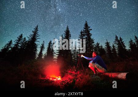 Photographe masculin faisant une séance photo dans les montagnes. Homme prenant des photos de nuit étoilée. Jeune homme assis sur un rondin à côté d'un feu de joie la nuit. Banque D'Images