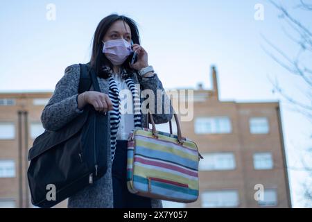 Femme d'affaires dans le masque chirurgical occupé à parler au téléphone tout en portant une mallette de documents et un sac à dos avec ordinateur de travail Banque D'Images