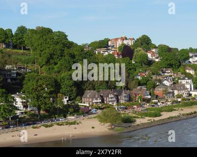 Vue côtière avec des maisons le long de la plage et une verdure luxuriante sur les collines sous un ciel clair, rive verte sur une rivière avec de nombreux arbres et tours, hambourg, Banque D'Images