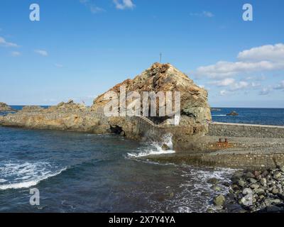 Une côte rocheuse avec un pont de pierre dans la mer et des vagues s'écrasant contre les rochers, plage de lave avec des montagnes et ciel bleu en arrière-plan, tener Banque D'Images