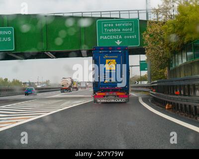 Un tronçon d'autoroute avec des camions voyageant sous la pluie, sous un passage supérieur avec des panneaux verts, jour de pluie sur l'autoroute A1, Piacenza, Italie, Europe Banque D'Images