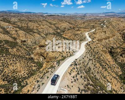 Une route solitaire mène à travers un terrain désertique aride et vallonné avec un fond rocheux escarpé et un ciel bleu, une vue aérienne, un camping-car voyageant le long d'un étroit ro Banque D'Images