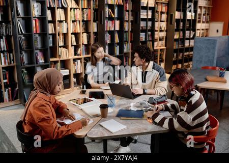 Vue en angle élevé de quatre étudiants masculins et féminins ethniquement divers assis dans la bibliothèque universitaire moderne faisant leurs devoirs Banque D'Images