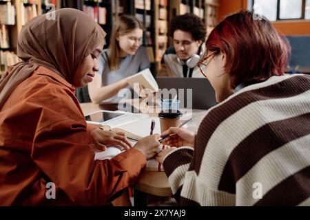 Deux filles diverses assises à table dans la bibliothèque de l'école moderne étudiant ensemble, prenant des notes dans un cahier Banque D'Images