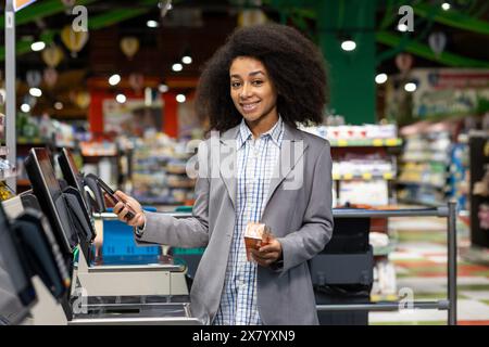 Dans une épicerie, une jeune femme qui utilise une machine à caisse automatique alors qu’elle tient un smartphone et un article d’épicerie. Cette scène représente une expérience d'achat moderne avec la technologie numérique Banque D'Images