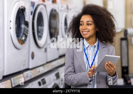 Dans un magasin d'électroménager, une femme heureuse aux cheveux bouclés sourit tenant une tablette. Elle compare les options devant les machines à laver et les sèche-linge, peut-être faire un achat ou recueillir des informations pour l'achat Banque D'Images