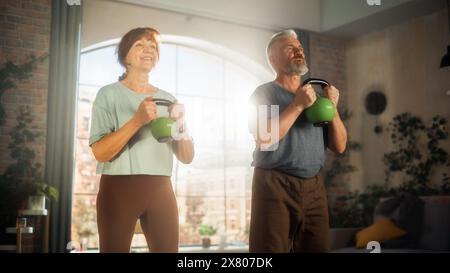 Heureux couple Senior faisant des exercices du matin et Kettlebell entraînement ensemble à la maison dans le salon ensoleillé. Concept de mode de vie sain, condition physique, loisirs, objectifs de couple, bien-être et retraite. Banque D'Images