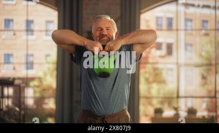 Strong Athletic Fit Senior homme soulevant une Kettlebell lourde, faisant des exercices de renforcement du noyau, gagnant de la masse musculaire pendant l'entraînement matinal à la maison dans Sunny Apartment. Concept de retraite et de remise en forme. Banque D'Images