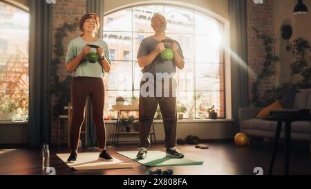 Couple aîné inspiré faisant des exercices matinaux et Kettlebell entraînement ensemble à la maison dans le salon lumineux. Concept de mode de vie sain, condition physique, loisirs, objectifs de couple, bien-être et retraite. Banque D'Images