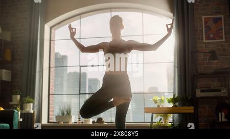 Jeune femme athlétique exerçant, pratiquant l'équilibre avec Vrikshasana Tree Yoga pose le matin dans son salon lumineux et ensoleillé. Mode de vie sain, Fitness, bien-être et concept de pleine conscience. Banque D'Images