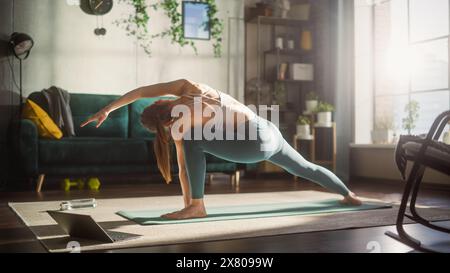 Jeune et attrayante femme athlétique exerçant, étirant et pratiquant le yoga avancé le matin dans le salon lumineux Sunny Home. Entraînement de fitness en ligne via un ordinateur portable. Banque D'Images