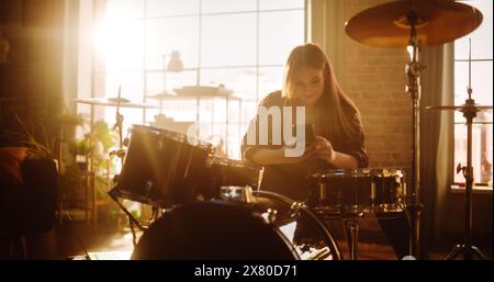 Jeune femme sort son smartphone pour répondre à un message sur les réseaux sociaux. Le batteur expressif a fini de jouer de la batterie dans un studio de répétition Loft Music rempli de lumière. Artiste musical a appris un Drum Solo. Banque D'Images