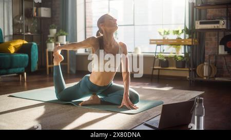 Jeune femme athlétique exerçant, étirant et pratiquant le yoga avancé le matin dans le salon lumineux Sunny Home. Entraînement de fitness en ligne via un ordinateur portable. Banque D'Images