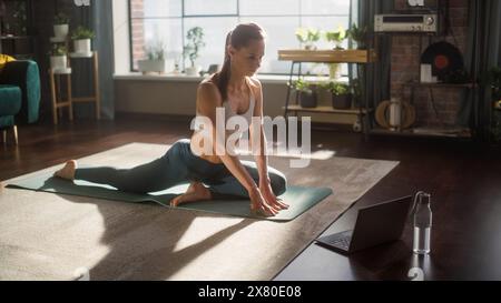 Athlétique jeune femme exerçant, étirant et faisant du yoga tout en regardant la vidéo d'entraînement sur un ordinateur portable. Belle femme en vêtements de sport pratiquant différentes poses Asana sur le tapis. Banque D'Images