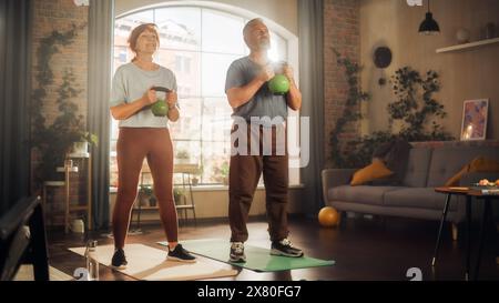 Heureux couple d'âge moyen faisant des exercices du matin et Kettlebell Workout ensemble à la maison dans le salon ensoleillé. Concept de mode de vie sain, condition physique, loisirs, objectifs de couple et retraite. Banque D'Images