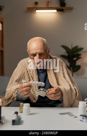 Un vieil homme souffrant de la grippe assis sur le canapé à la maison tient des pilules dans ses mains. Concept de maladies saisonnières et de virus Banque D'Images