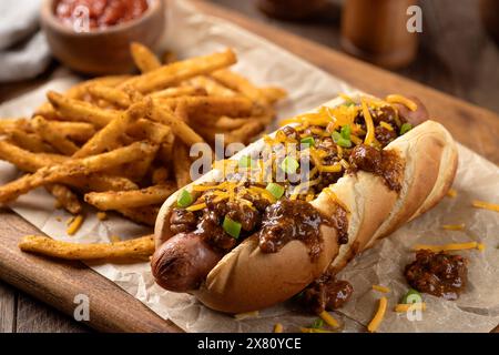 Chili hot dog avec fromage cheddar râpé et frites assaisonnées sur une planche de bois Banque D'Images