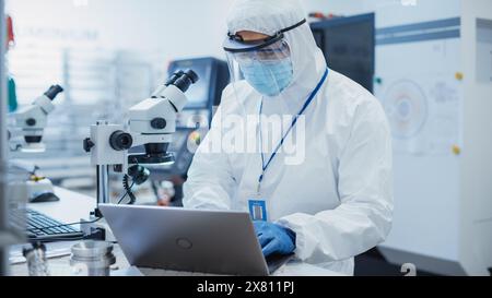 Usine de fabrication électronique : ingénieur en combinaison stérile travaillant sur un ordinateur portable, examinant une carte de circuit imprimé avec des micropuces à travers un microscope et testant de nouveaux équipements électriques. Banque D'Images