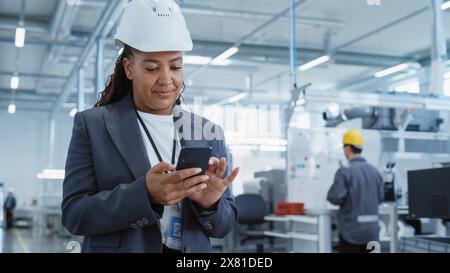 Portrait d'une ingénieure noire en casque de sécurité debout et à l'aide d'un smartphone à l'usine de fabrication d'électronique. Le technicien rédige un message et vérifie son horaire. Banque D'Images