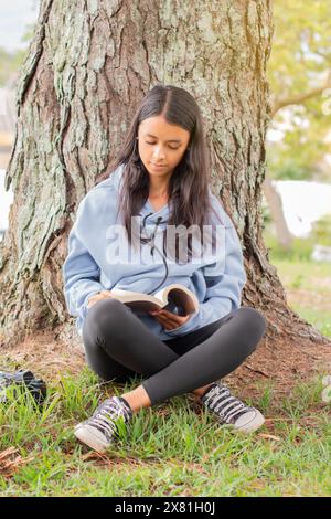 style de vie : jeune latina lisant un livre assis dans l'herbe à côté d'un arbre Banque D'Images