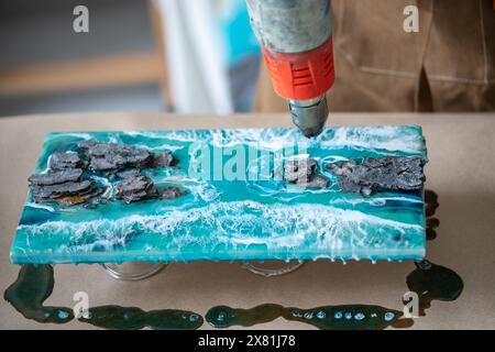 Artiste travaillant avec de la résine époxy liquide, créant la mer sur un panneau de résine en deux parties, soufflant le séchoir thermique Banque D'Images