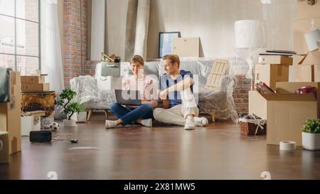 Heureux jeunes propriétaires : charmant couple assis sur le sol de l'appartement nouvellement acheté utiliser l'ordinateur portable. E-commerce achats en ligne. Prêt hypothécaire, investissement immobilier, Sweet Home. Grand angle Banque D'Images