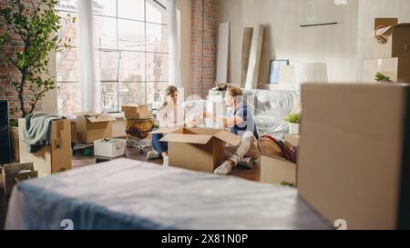 Heureux propriétaires emménageant : beau couple assis sur le sol de Cozy Apartment déballage des boîtes en carton. Prêt hypothécaire, immobilier, Sweet Home pour jeune famille. Journée lumineuse pleine de souvenirs heureux Banque D'Images