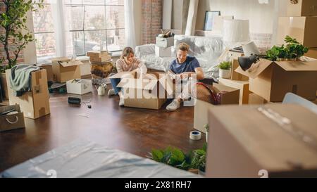 Heureux propriétaires emménageant : beau couple assis sur le sol de Cozy Apartment déballage des boîtes en carton. Prêt hypothécaire, immobilier, Sweet Home pour jeune famille. Journée lumineuse pleine de souvenirs heureux Banque D'Images