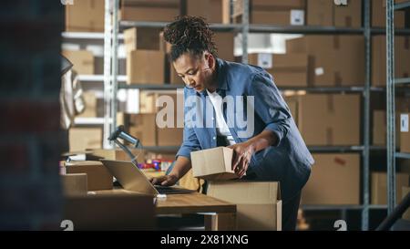 Gestionnaire d'inventaire préparant un petit colis de carton pour l'affranchissement. Propriétaire multiethnique afro-américaine de petite entreprise travaillant sur un ordinateur portable dans un entrepôt. Banque D'Images