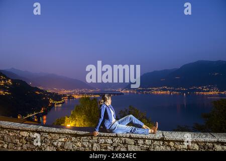 Femme assise sur un mur avec vue panoramique sur le lac majeur et la montagne au crépuscule à Ronco sopra Ascona, Tessin, Suisse. Banque D'Images
