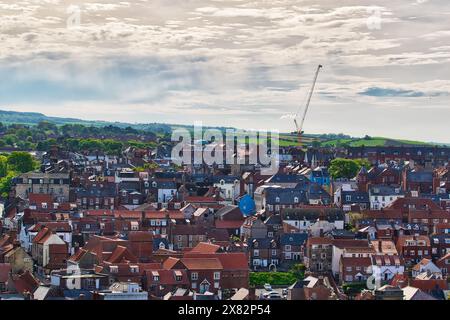 Vue aérienne d'une petite ville avec des maisons au toit rouge et une grue en arrière-plan. La ville est entourée de champs verdoyants et de collines sous un ciel nuageux. Banque D'Images