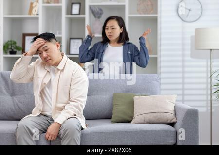 Un couple stressé se disputant à la maison dans un salon moderne. L'homme est assis sur le canapé, tandis que la femme se tient derrière les bras levés. Banque D'Images