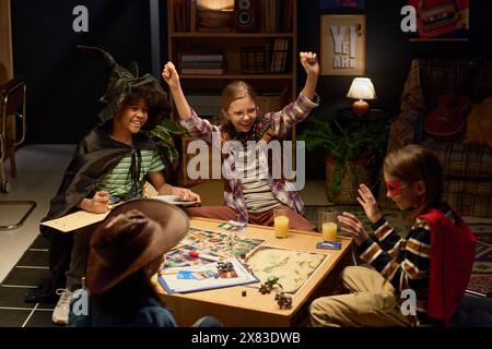 Deux enfants extatiques en costumes de leurs personnages levant la main et exprimant le triomphe de la victoire dans le jeu de table avec des amis Banque D'Images