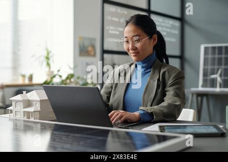 Employé féminin asiatique souriant regardant l'écran d'ordinateur portable et tapant tout en étant assis par le lieu de travail avec modèle de maison et panneau solaire Banque D'Images