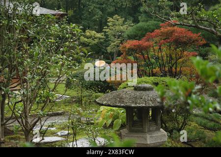 Un jardin japonais serein avec une lanterne en pierre, une végétation luxuriante et une personne portant un chapeau traditionnel. Le jardin dispose de diverses plantes, y compris tr Banque D'Images