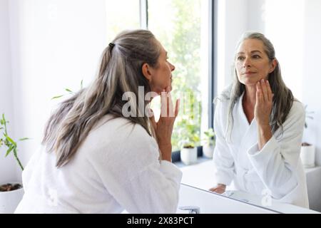 À la salle de bain à la maison, femme caucasienne mature portant une robe blanche, examinant son visage dans un miroir Banque D'Images